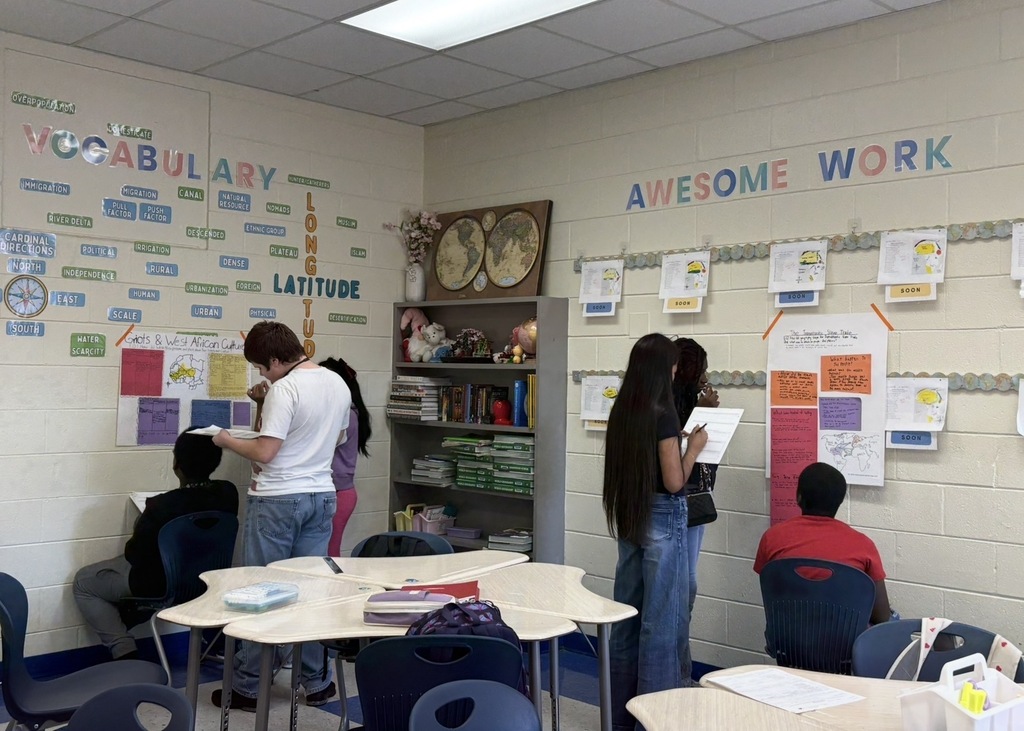 7th grade social studies students are conducting a gallery walk by viewing different student group posters that share information about West African culture as they wrapped up this unit in class. Students are shown walking around with a paper and pencil in hand as they look at the different posters taped on the wall in different places in the classroom.