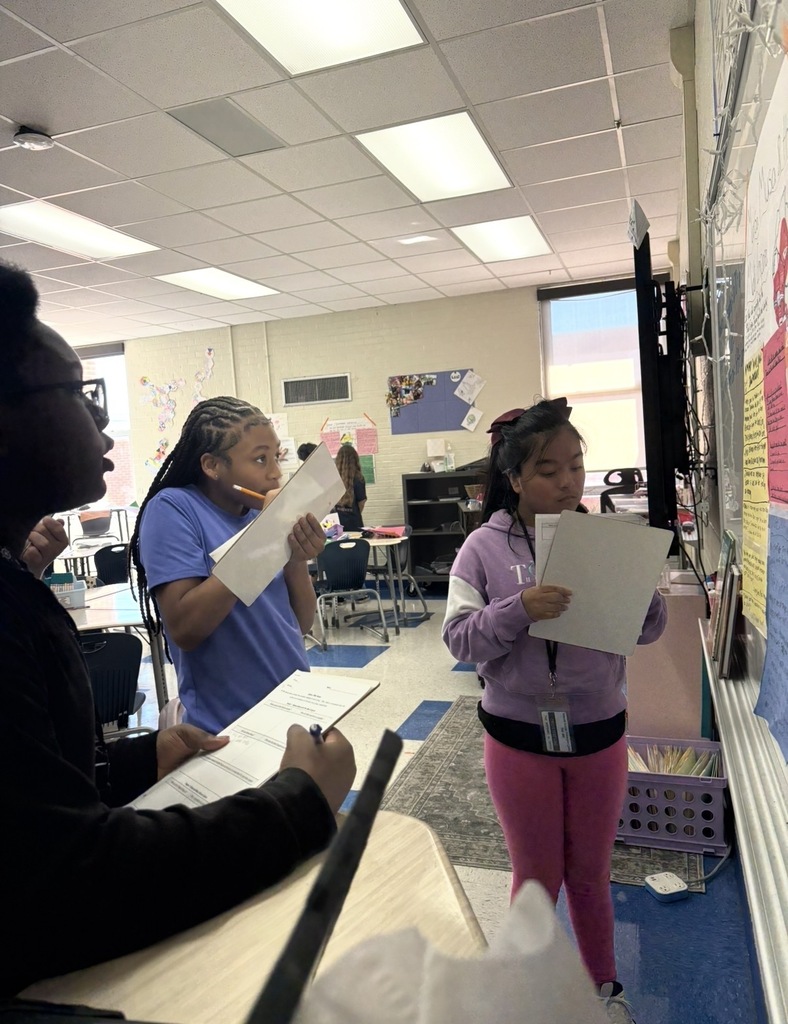 7th grade social studies students are conducting a gallery walk by viewing different student group posters that share information about West African culture as they wrapped up this unit in class. Students are shown walking around with a paper and pencil in hand as they look at the different posters taped on the wall in different places in the classroom.