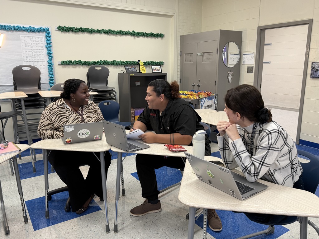 Dr. Johnson is shown talking with two 7th grade teachers while all three of them are seated in student desks next to one another.