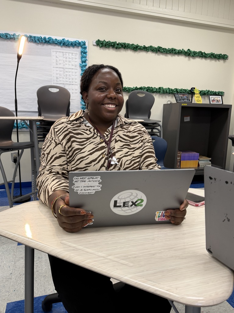 Dr. Johnson is shown sitting at a student desk with her hands on the monitor of her laptop and smiling at the camera.