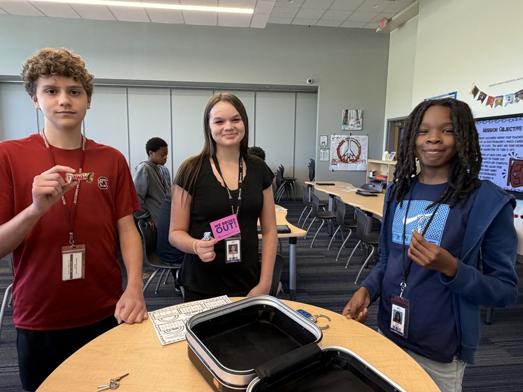 A group of three students are shown standing at the open lock box that they broke into with the correct code. They are all looking at the camera smiling and holding either a piece of candy that was their prize, the sign that states, "We escaped!," or the answer sheet they recorded their answers to the questions and puzzles on.