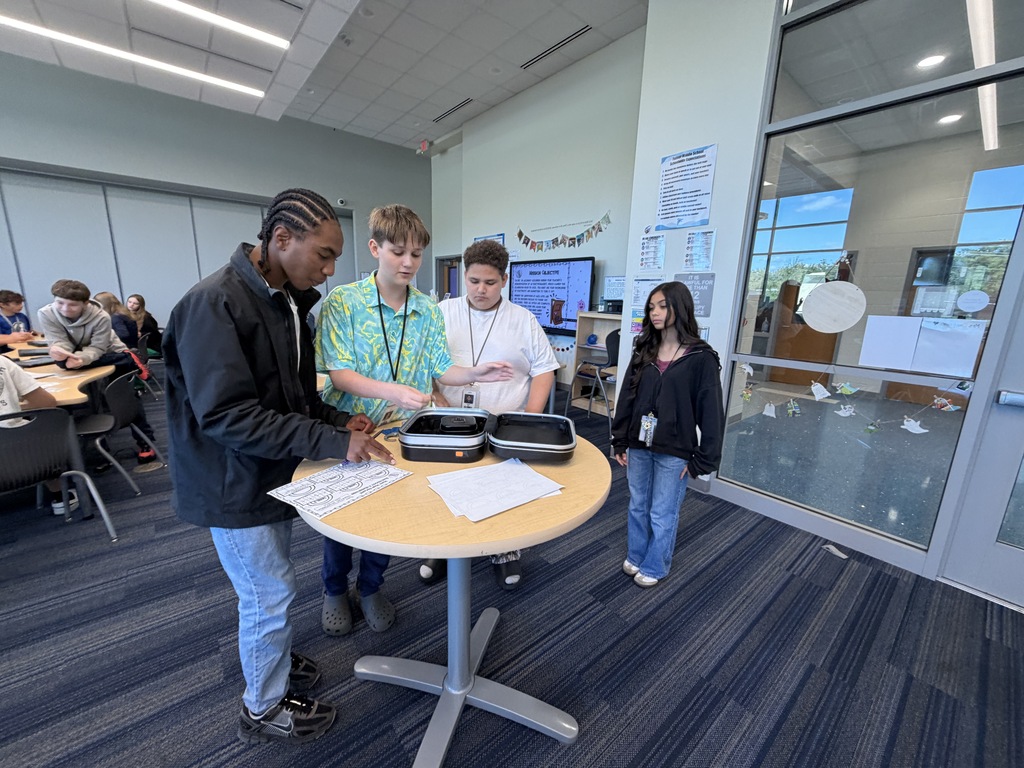 A group of students are standing around a tall table as they work to enter the four digit code into a combination lock to break into the lockbox and retrieve their price since they were the first group to successfully complete all five levels of the science review.