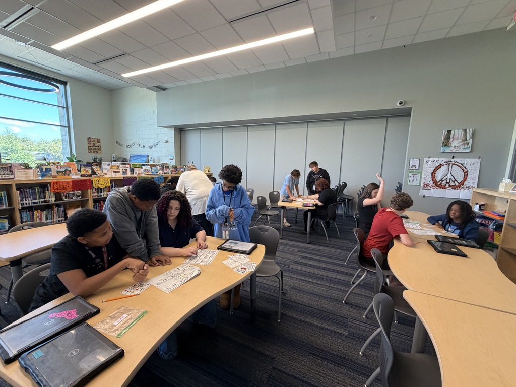 The class of students are shown sitting or standing tables  working together on answering questions related to their electricity and magnetism unit in 8th grade science class.
