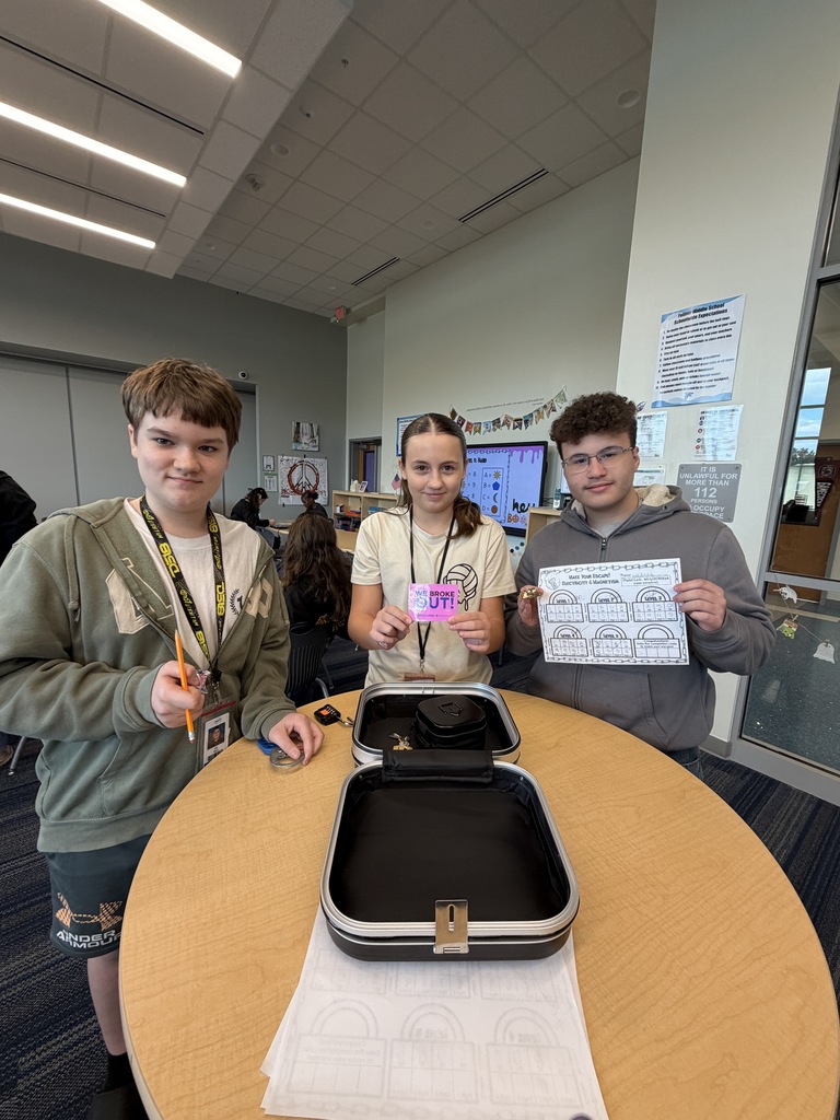 A group of three students are shown standing at the open lock box that they broke into with the correct code. They are all looking at the camera smiling and holding either a piece of candy that was their prize, the sign that states, "We escaped!," or the answer sheet they recorded their answers to the questions and puzzles on.