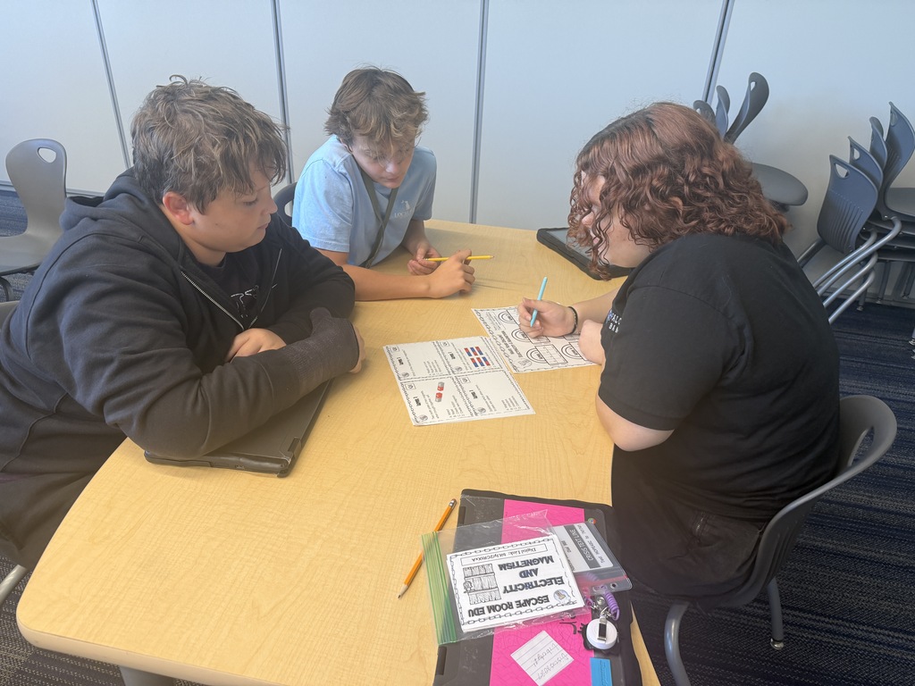 Three students are seated at a table working together on answering questions related to their electricity and magnetism unit in 8th grade science class.