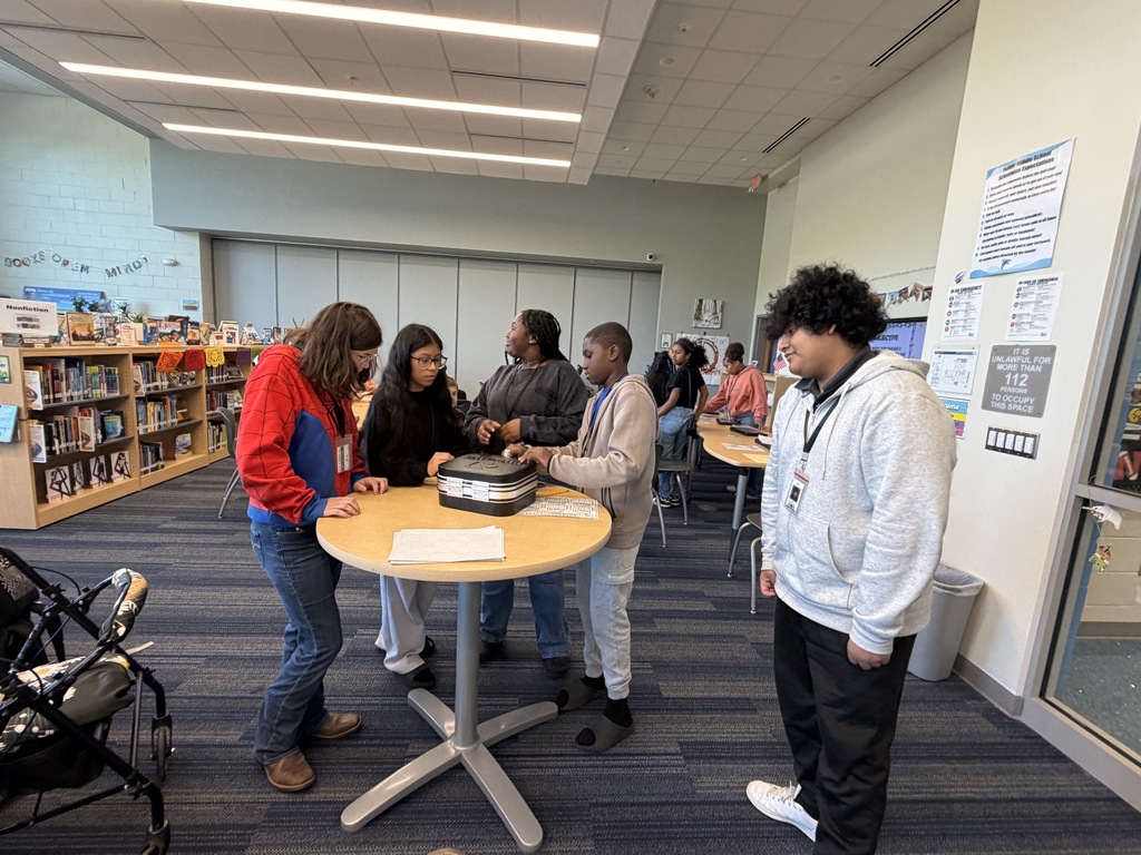 A group of students are standing around a tall table as they work to enter the four digit code into a combination lock to break into the lockbox and retrieve their price since they were the first group to successfully complete all five levels of the science review.