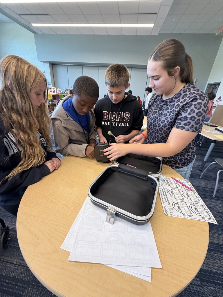 A group of students are standing around a tall table as they work to enter the four digit code into a combination lock to break into the lockbox and retrieve their price since they were the first group to successfully complete all five levels of the science review.