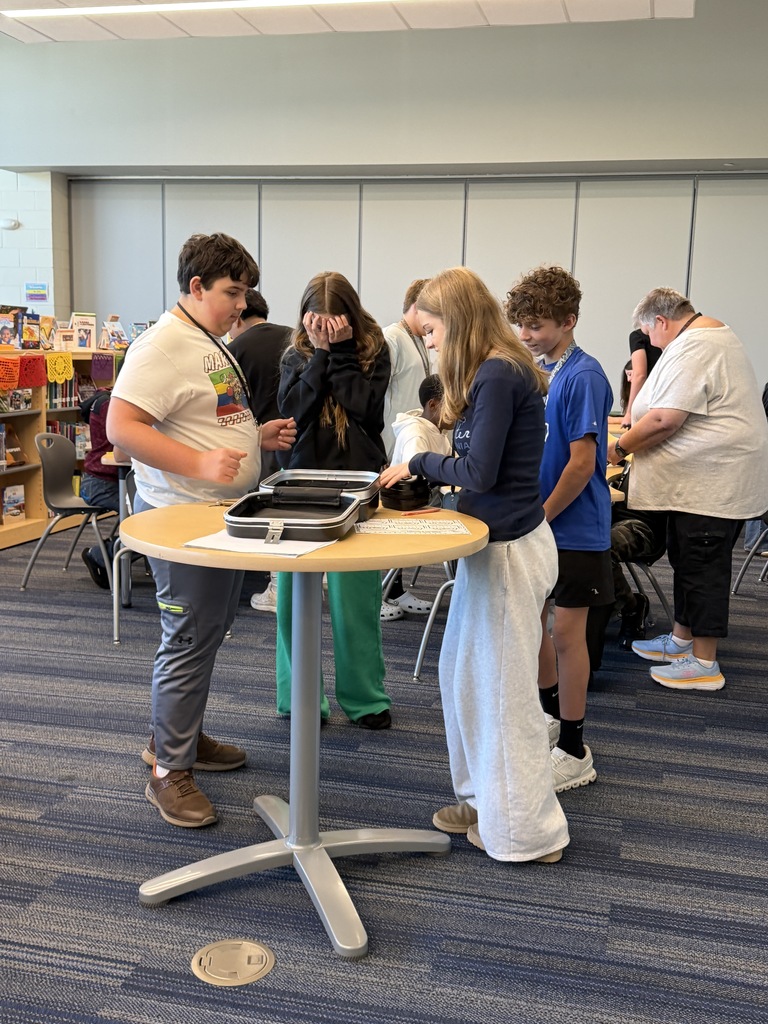 A group of students are standing around a tall table as they work to enter the four digit code into a combination lock to break into the lockbox and retrieve their price since they were the first group to successfully complete all five levels of the science review.