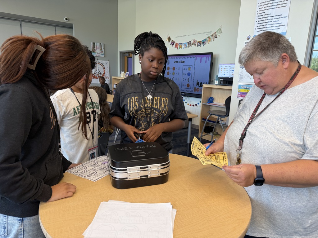 A group of students are standing around a tall table as they work to enter the four digit code into a combination lock to break into the lockbox and retrieve their price since they were the first group to successfully complete all five levels of the science review. The science teacher is also standing at the table and looking at her answer key for the answers to the questions and puzzles.
