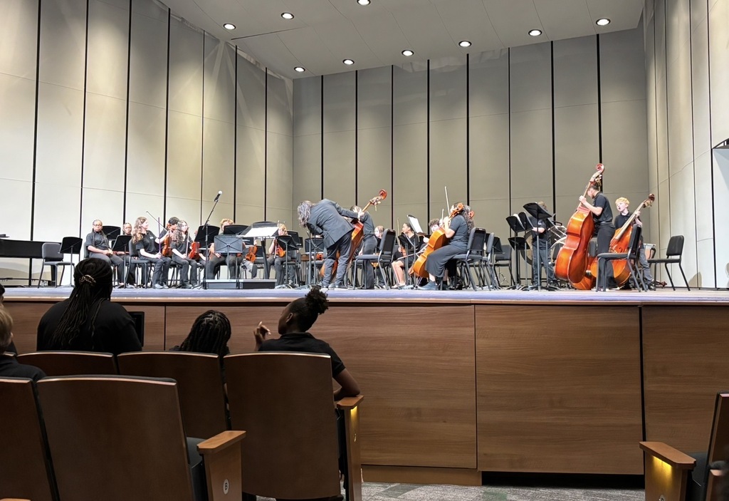 The orchestra director is bent slightly to the side on the front middle of the stage fixing a cello. Other orchestra students are seated holding their instruments.
