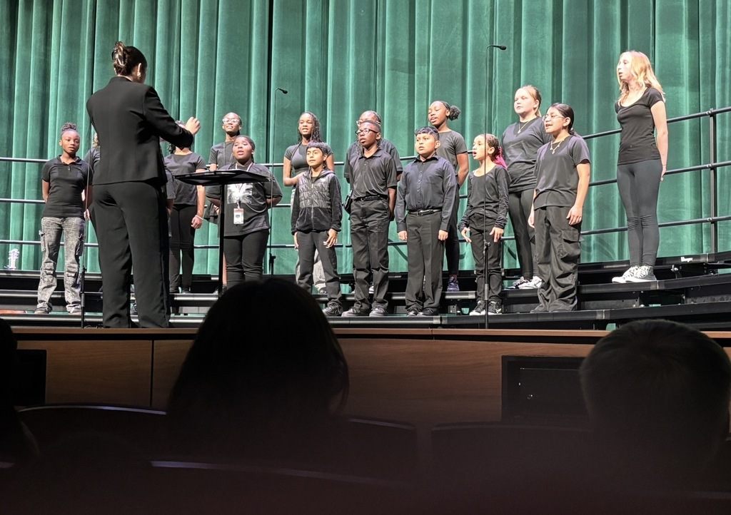The choir director is shown at the front middle of the stage facing the 6th grade chorus students who are standing on the rafter. She is looking at the students and has her arms slightly up directing them.