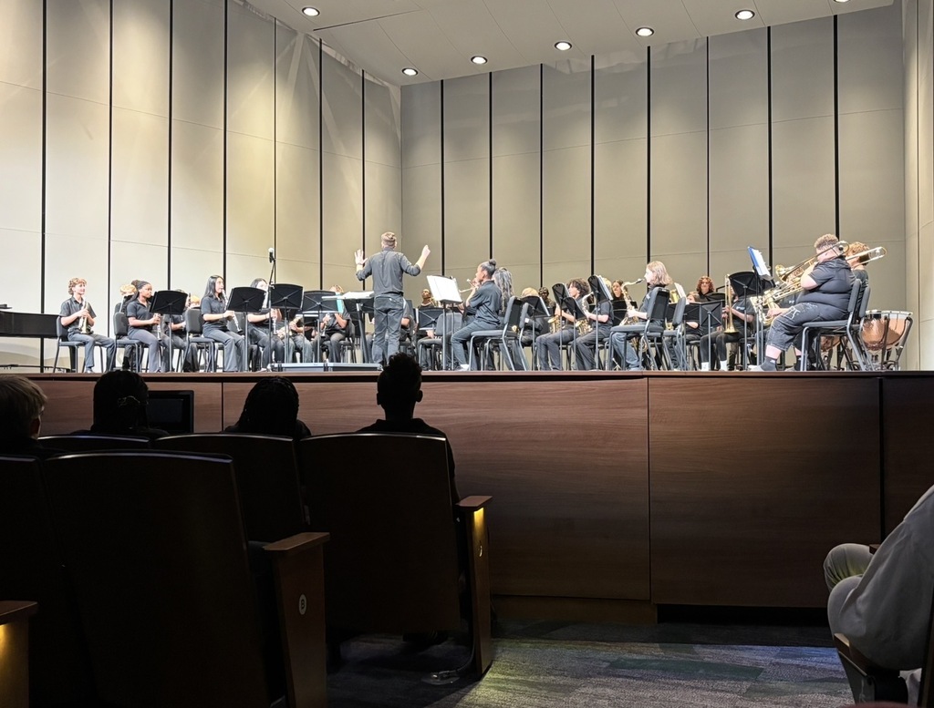 The band director is standing towards the front middle of the stage and has his hands half way up conducting his band students in their Fall Fine Arts Concert.