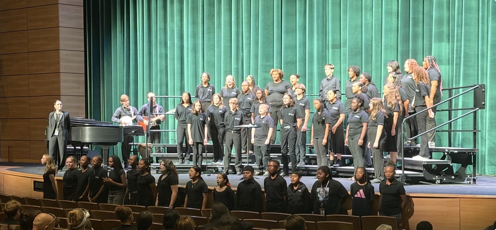 6th, 7th, and 8th grade Chorus students are standing on rafters or on the floor in front of the stage for a Fall Fine Arts Concert. The Choir Director is standing to the side of the stage in front of a piano. Two guitar players are standing next to the rafters.