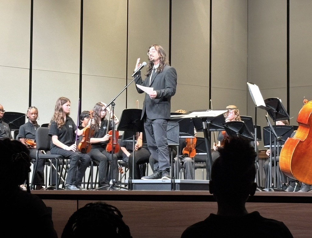 The orchestra director is shown facing the crowd on a stage and talking into the microphone holding a piece of paper. Some of the orchestra students are shown in the chairs on stage holding their instruments.