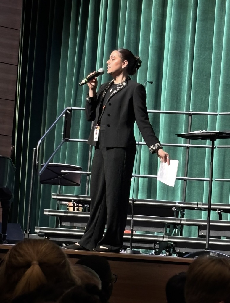 The choir director is shown on stage facing the crowd with a microphone in her hand talking. She holds a piece of paper downward in her left hand.