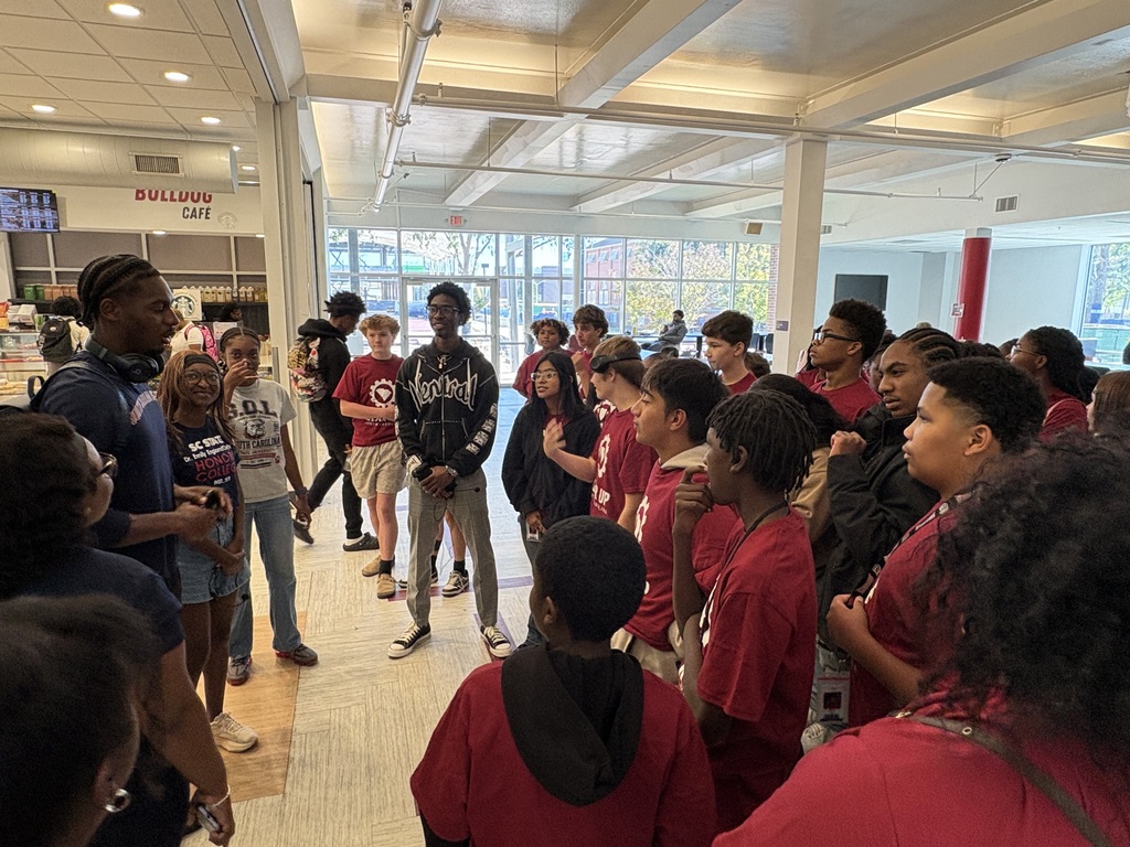 A group of male and female students are standing inside of a building listening to one of the adult tour guides.