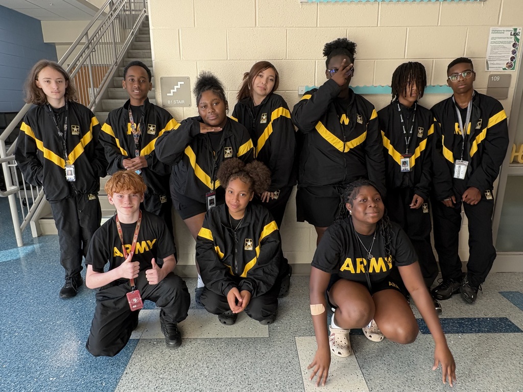 A group of 8th grade JROTC students are standing in a row and three of them are squatting downwards in front of those standing. These students are dressed in their Physical Training uniforms to do PT during their JROTC class.