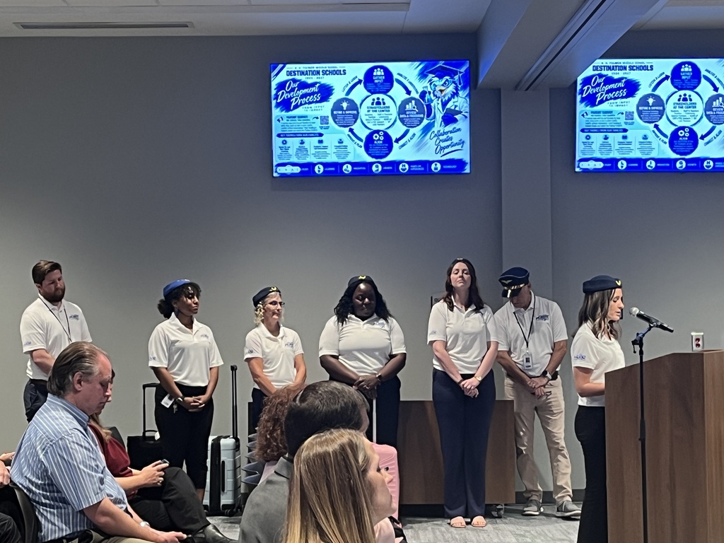 FMS faculty members and administration team are standing near a wall during the board meeting. One staff member is standing at the podium speaking into the microphone. There are two TVs posted on the wall with part of the presentation.