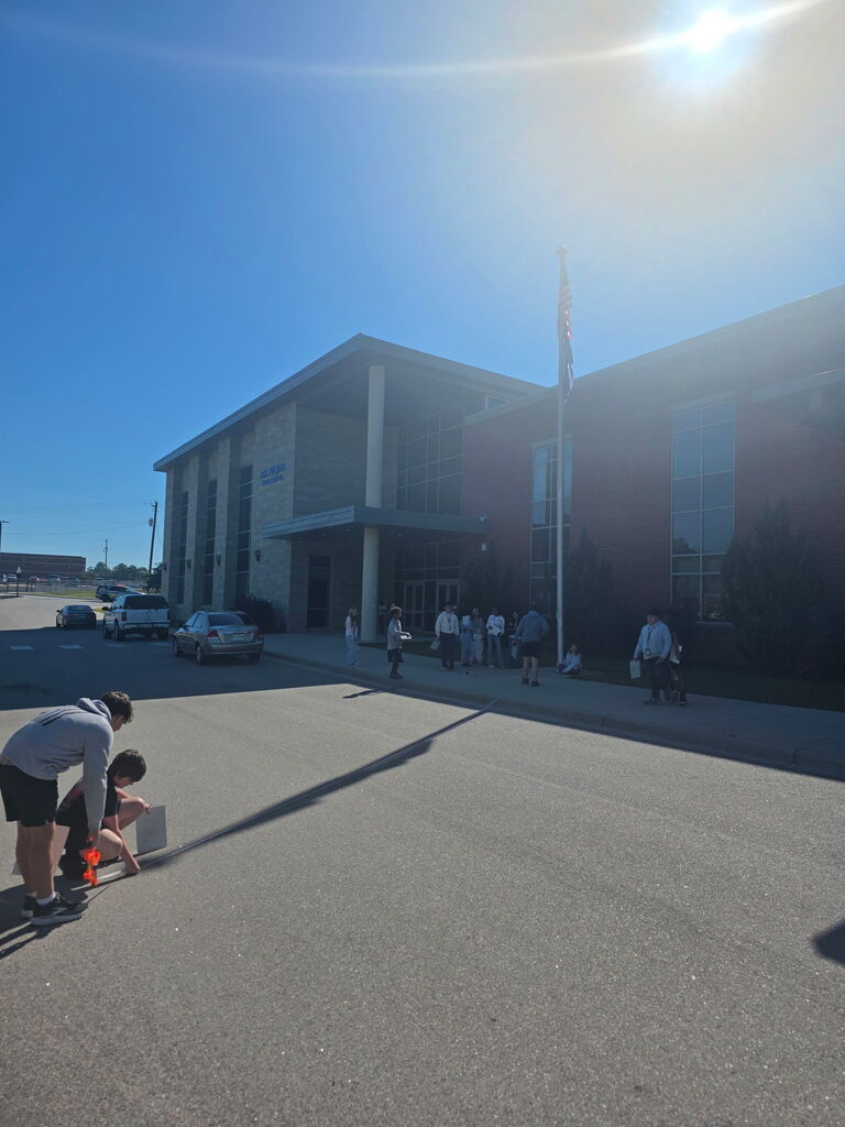 Two students are in the road parking lot squatting down to measure the flag pole's shadow.