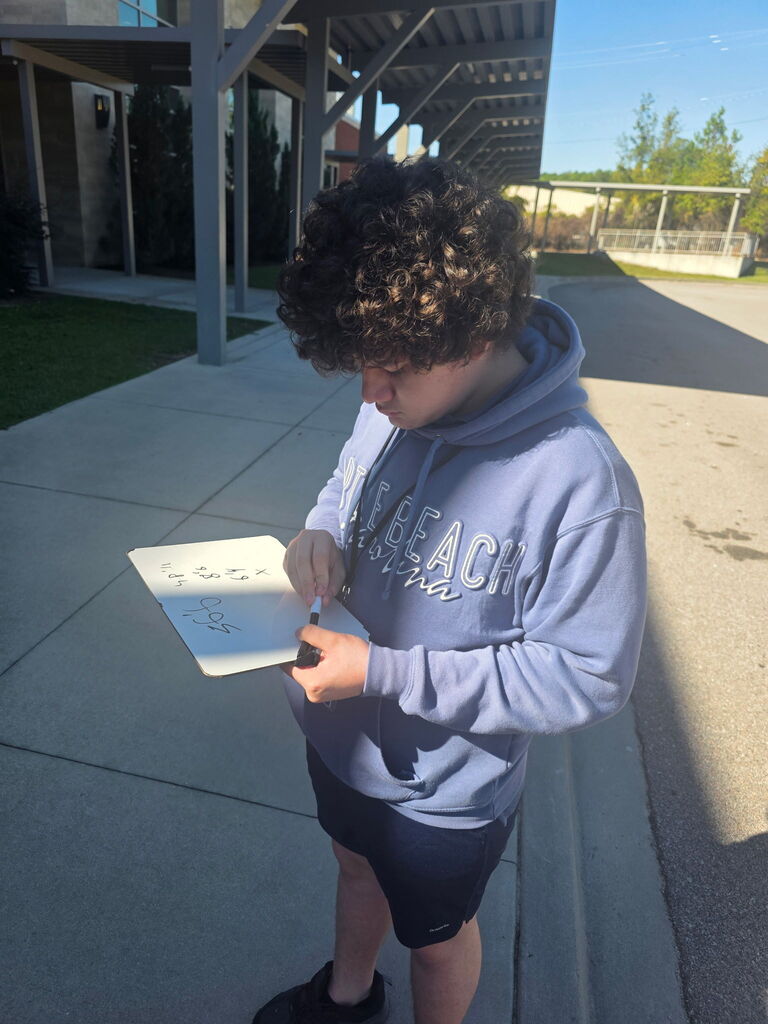 A student is shown standing outside on the sidewalk recording numbers on his dry erase board that he is holding.
