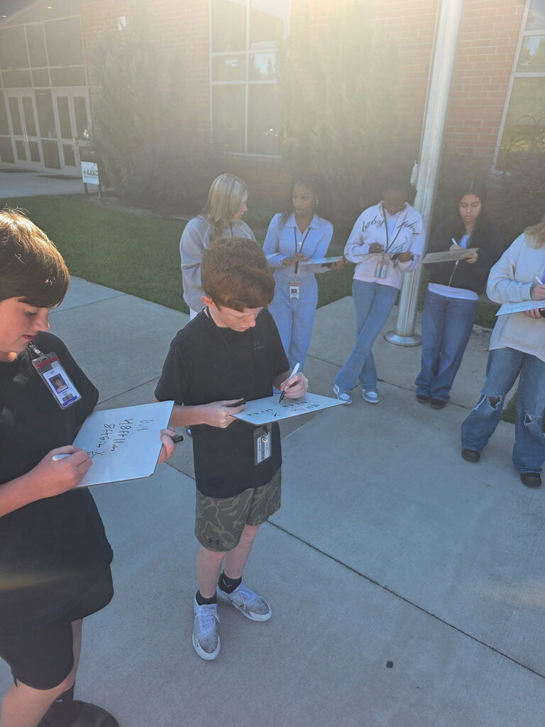 A group of students are shown standing outside of the school on the sidewalk near the flagpole recording measurements on an individual dry erase board.