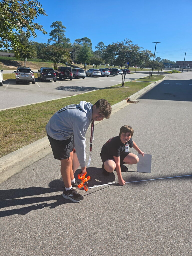 Two students are shown looking at the camera as they use a measuring tape tool to measure the flagpoles shadow on the ground.