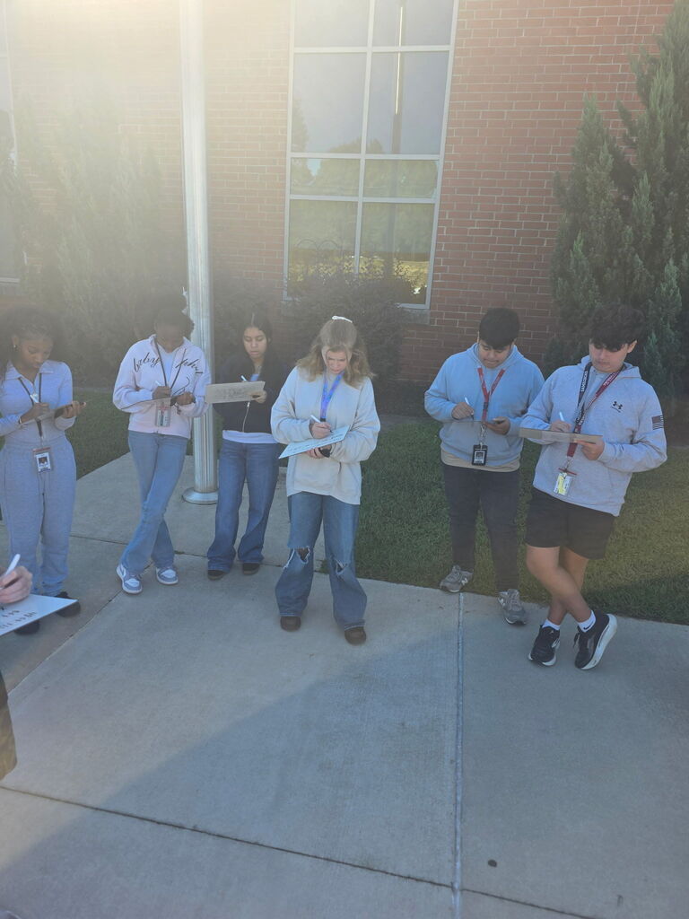 A group of students are shown standing outside of the school on the sidewalk near the flagpole recording measurements on an individual dry erase board.