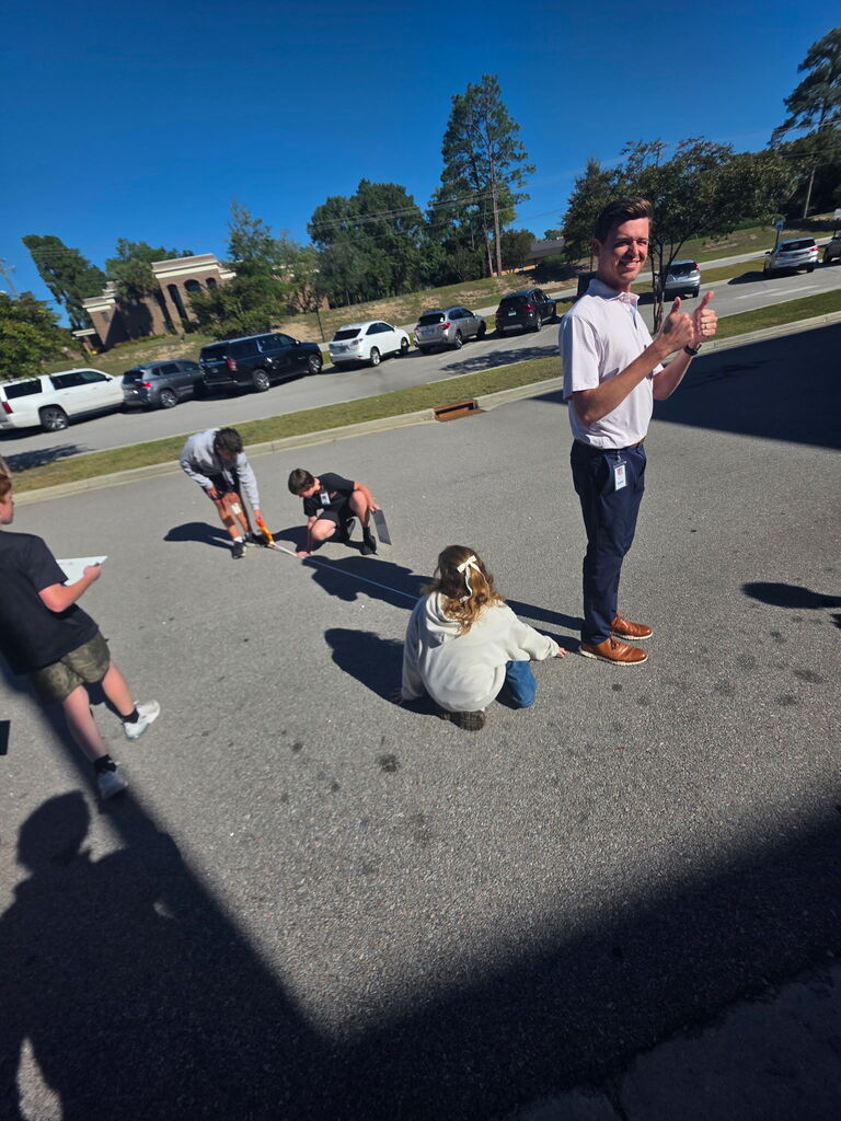 Mr. Boland is shown standing in the parking lot road as students are measuring his shadow on the ground. Three students are shown squatting on the ground to do the measuring and holding of hte tool. One student is shown standing and walking towards the two students looking at the number on the measuring tape at the top of Mr. Boland's shadow.