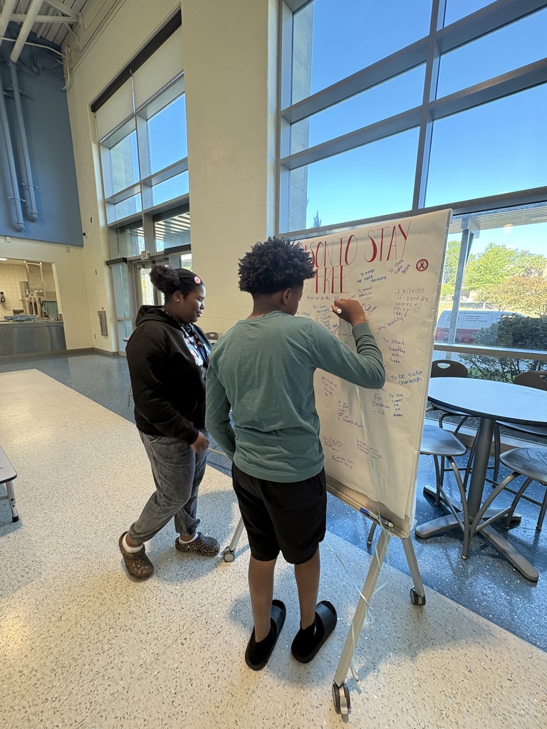 Two students are standing at a board on wheels in the cafeteria. They are signing their names and making a pledge to stay drug free. The poster at the top has red large, bold letters stating, "My Pledge to Stay Drug Free..."