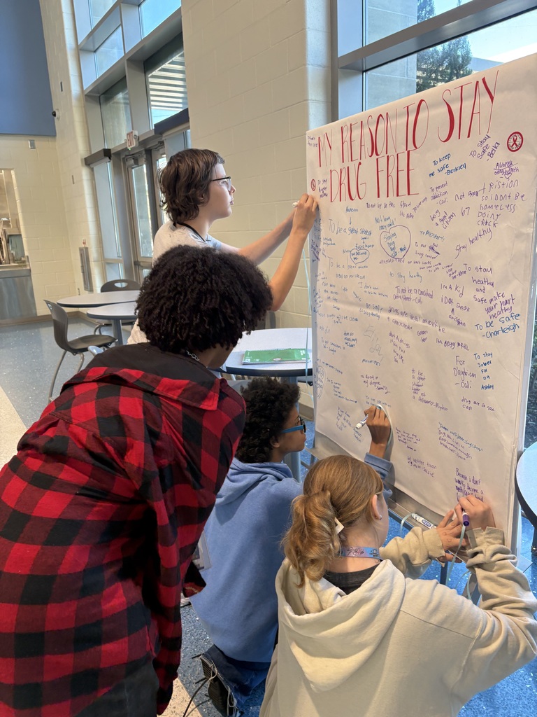 A group of students are standing at a board on wheels in the cafeteria. They are signing their names and making a pledge to stay drug free. The poster at the top has red large, bold letters stating, "My Pledge to Stay Drug Free..."