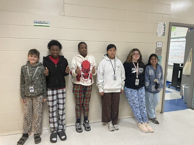 A group of 6th grades are standing lined up against the wall in the hallway wearing pajama pants for Red Ribbon Week. Two of the boys in the photo are showing two thumbs upo.