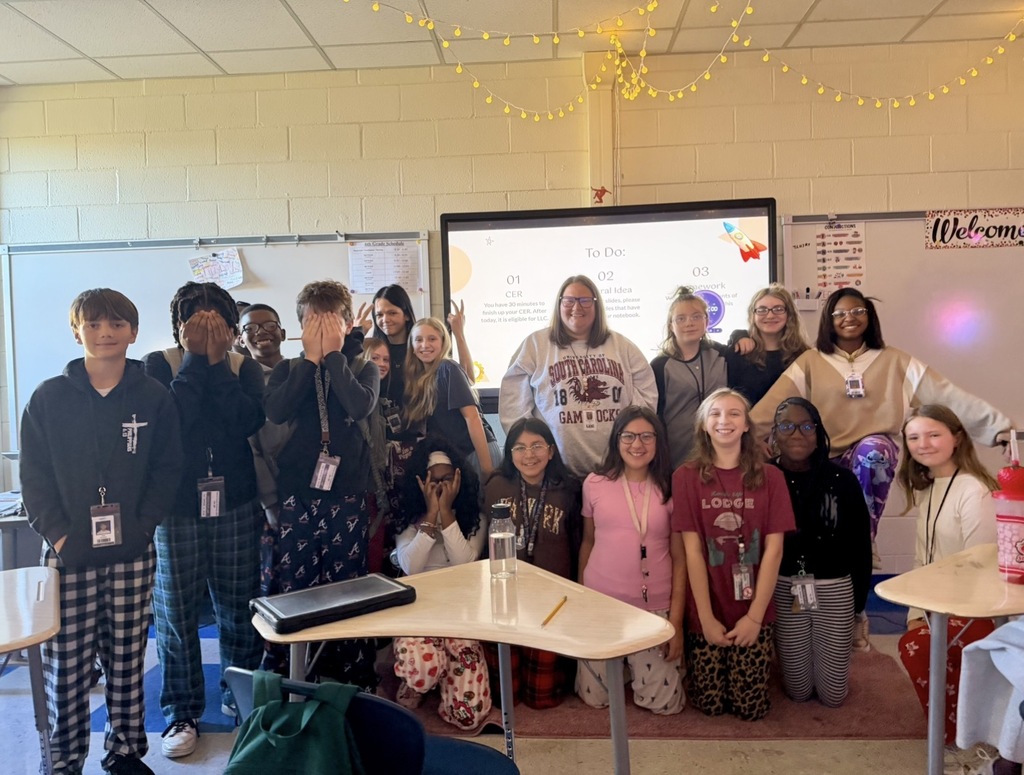 A group of students and their teacher are standing in front of the board smiling at the camera wearing pajama pants.