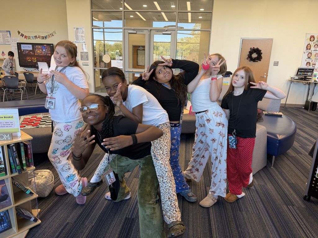 A group of female students are standing in the library making silly poses and looking at the camera wearing pajama pants.