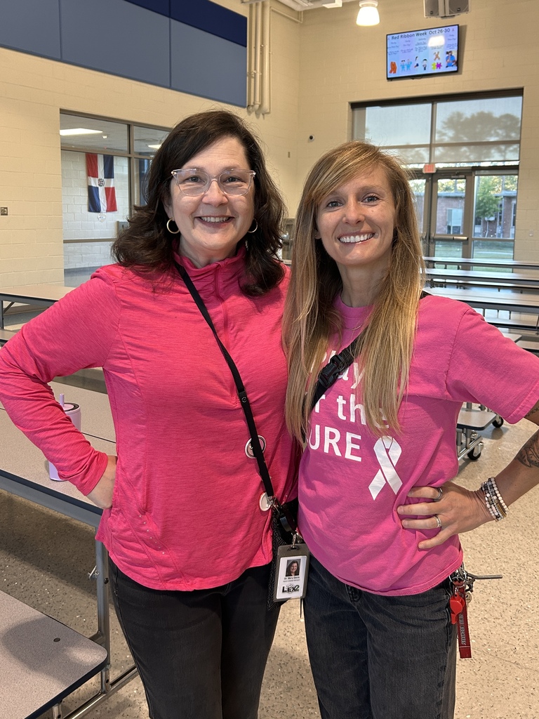 Two faculty members are standing in the hallway side by side smiling at the camera wearing pink for Breast Cancer Awareness.