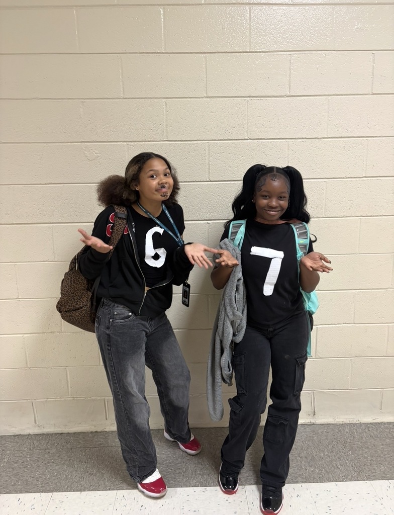 Two students stand side by side smiling at the camera wearing the exact clothing attire for Twin Day at school. They are both holding their arms and hands in a half ben outward position to match the hand motion for the common phrase "6-7" because one student has a white letter 6 on her shirt and the other has the white letter 7 on her shirt.