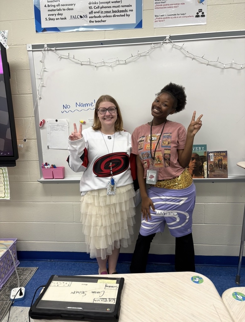 A teacher and a student stand together smiling in the front of a classroom both holding a peace sign and wearing mismatched clothing.