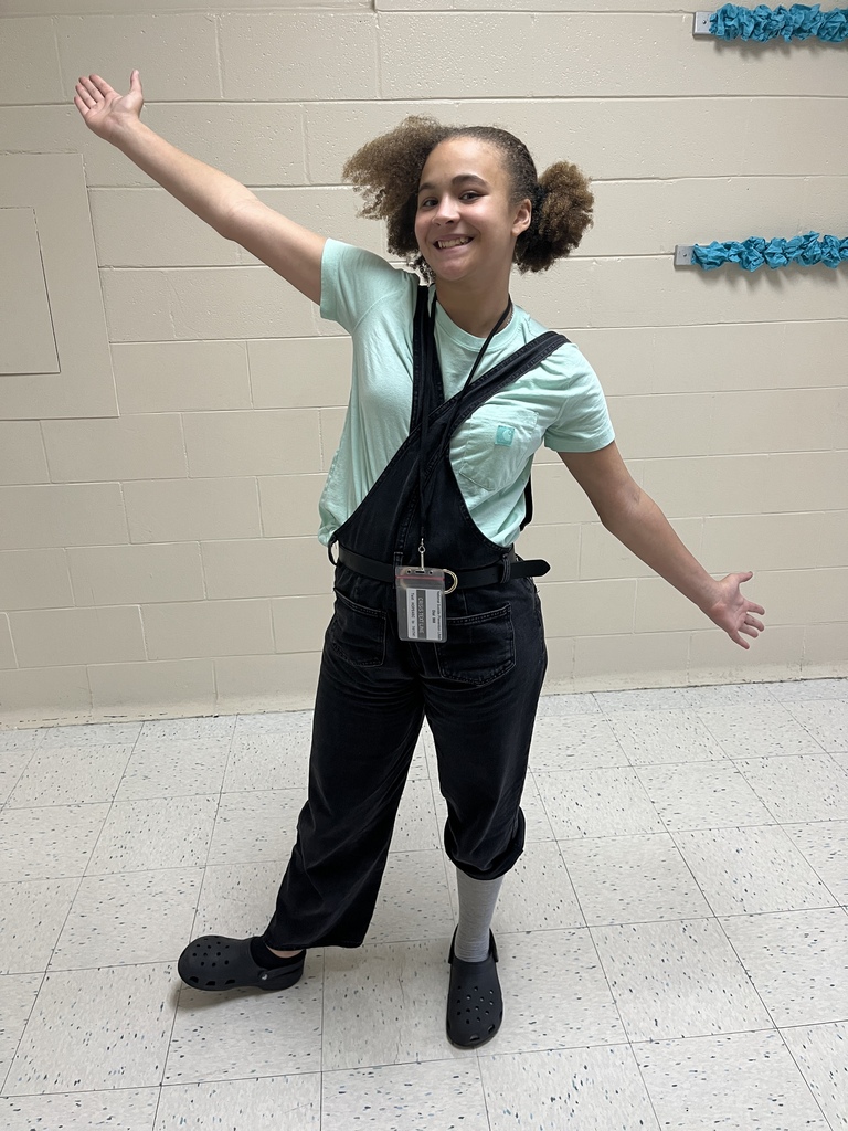 One student stands int he hallway with arms outstretched and smiling wearing overalls backwards and one hair in a pig tail poof and the other half of her side of her hair not in a pig tail.