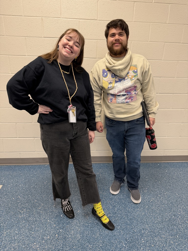 Two faculty member stand side by side smiling at the camera wearing mismatched clothing and shoes.