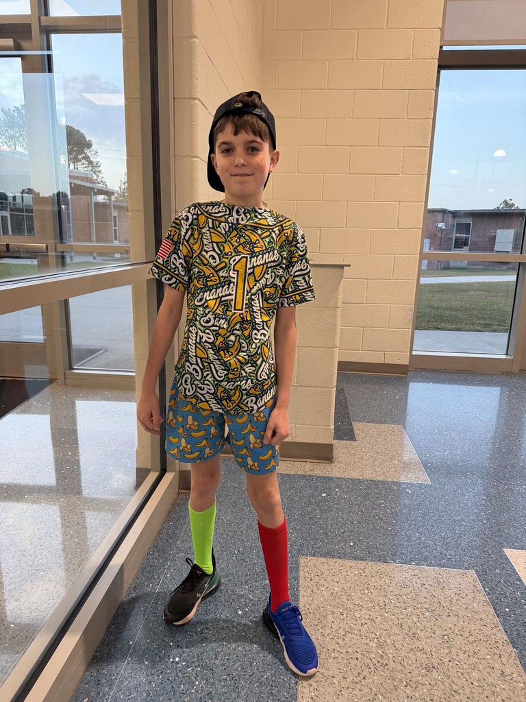 A student stands in the cafeteria smiling at the camera with a mismatched banana shirt, shorts, different shoes, and different colored socks. He also has a ball cap on his head that is turned backwards.