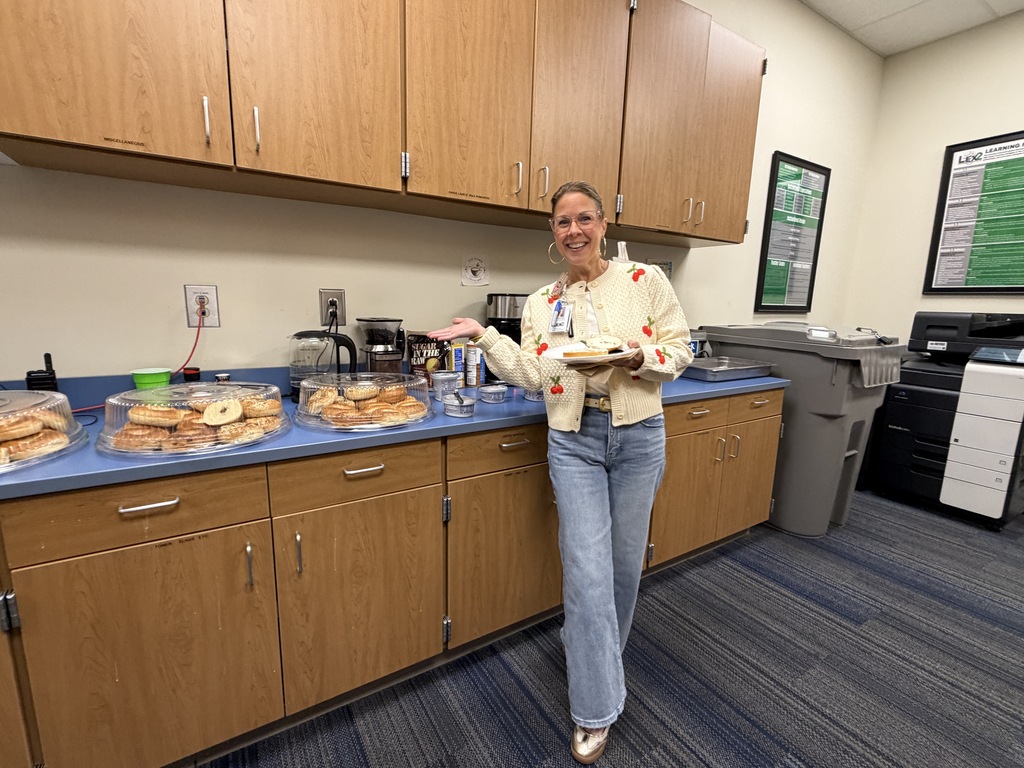 Nurse McCurdy stands in front of the bagel buffet holding a plate with a bagel on it in her hand and the other hand stretched out showing the bagels. She's smiling at the camera.