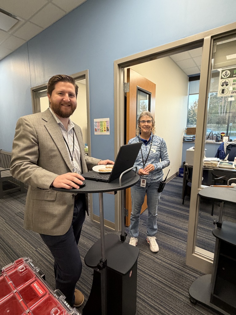 Mr. Ebener and Ms. Norris are standing in the hallway in the morning outside of their offices. Mr. Ebener has his hand on his computer mouse and on the tall rolling desk. Mr. Ebener has a bagel on a plate on top of his laptop. They are both smiling at the camera.
