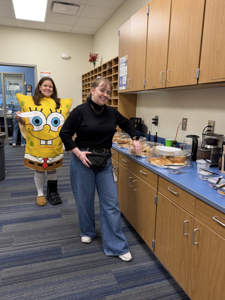 Ms. Skeen and Mrs. Shearer stand at the bagel bar getting bagels. They are smiling at the camera.