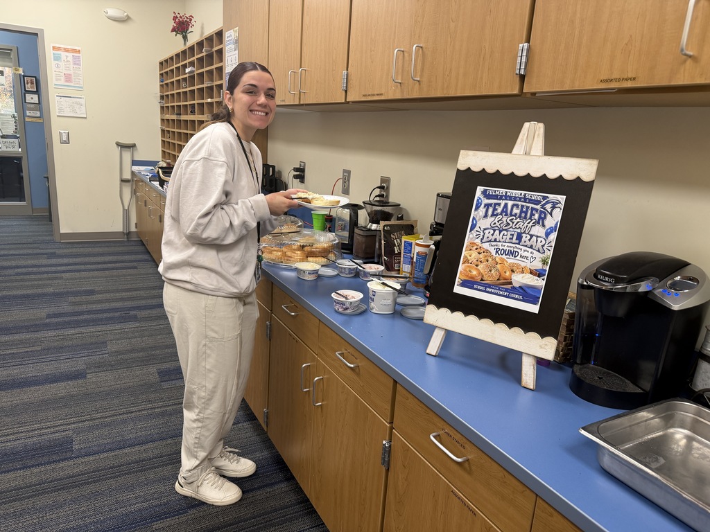 Ms. De Sena is standing at the bagel bar holding her plate with a bagel on it and smiling at the camera.