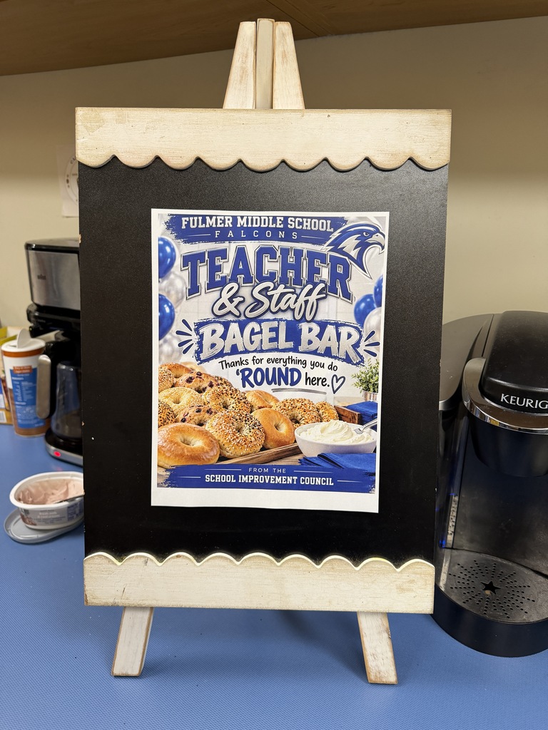 A picture of a sign taped to a black chalkboard like easel. The sign states Fulmer Middle School Falcons Teacher and Staff Bagel Bar Thanks for everything you do 'round here." from the School Improvement Council. next to the bagel bar. This sign is in blue and white and has a photo of a platter of bagels and a bowl of white cream cheese.