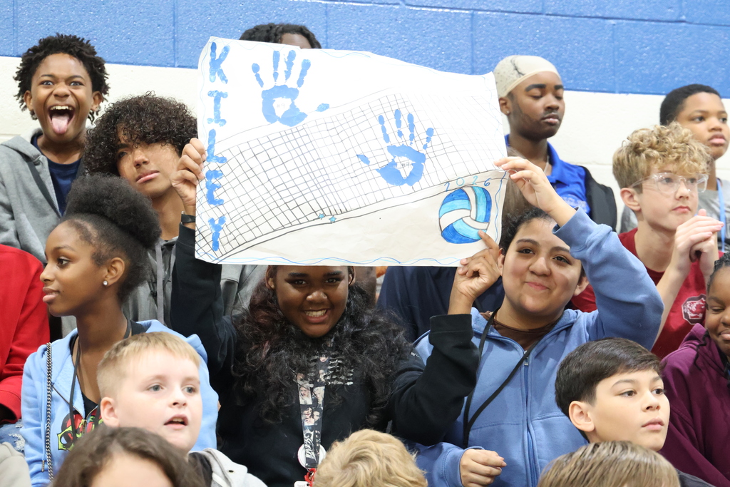 Students are shown sitting in the crowd. Some are smiling, some are looking off into the distance, one is looking at the camera with his tongue out, and two girls are smiling while holding up a poster they made with the name "Kiley" written on the left side, two blue handprints, a volleyball and a volleyball net.