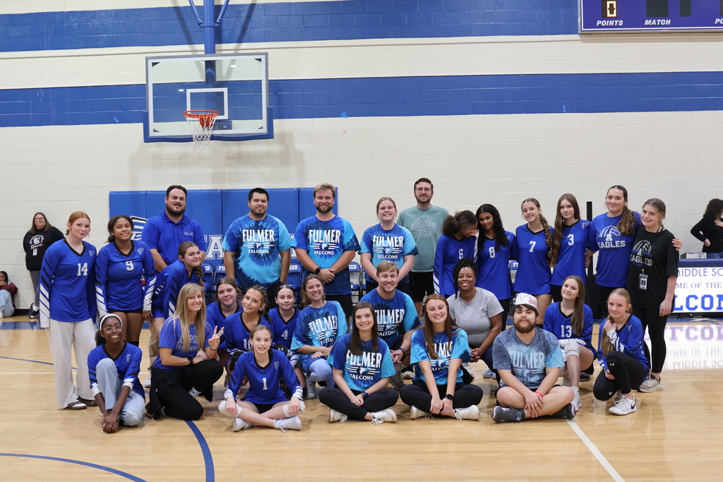A group photo of the lady falcon volleyball players with some of Fulmer's faculty and staff who participated in the volleyball game.  They are standing and sitting down smiling at the camera in the gym.