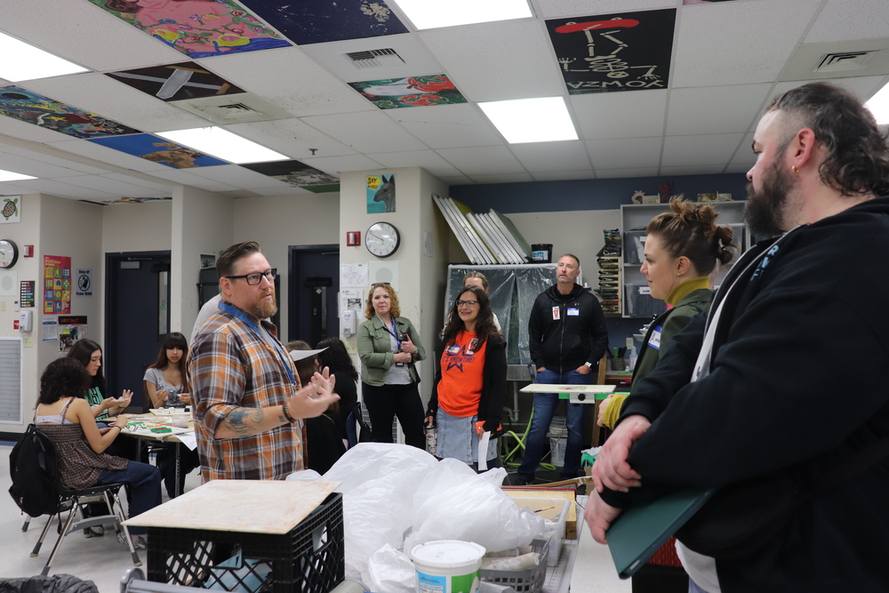 man in plaid shirt talks to a group of adults in a school classroom