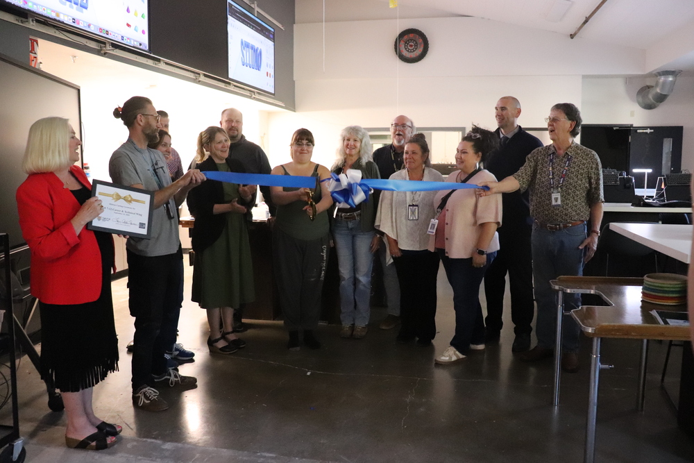 Men and women stand in a classroom with the woman in the center holding large scissors preparing to cut a long blue ribbon