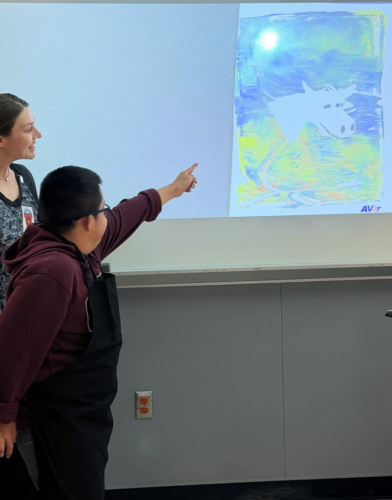 In this image, the student displays his artwork, pointing to the abstract art with an image in the center of it.