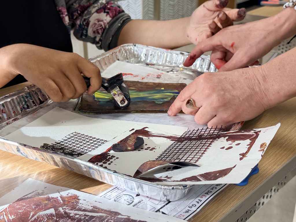 In this image the Sitka K-8 Create instructor helps a student hold their artwork as they roll over to press the ink/paint onto their paper.